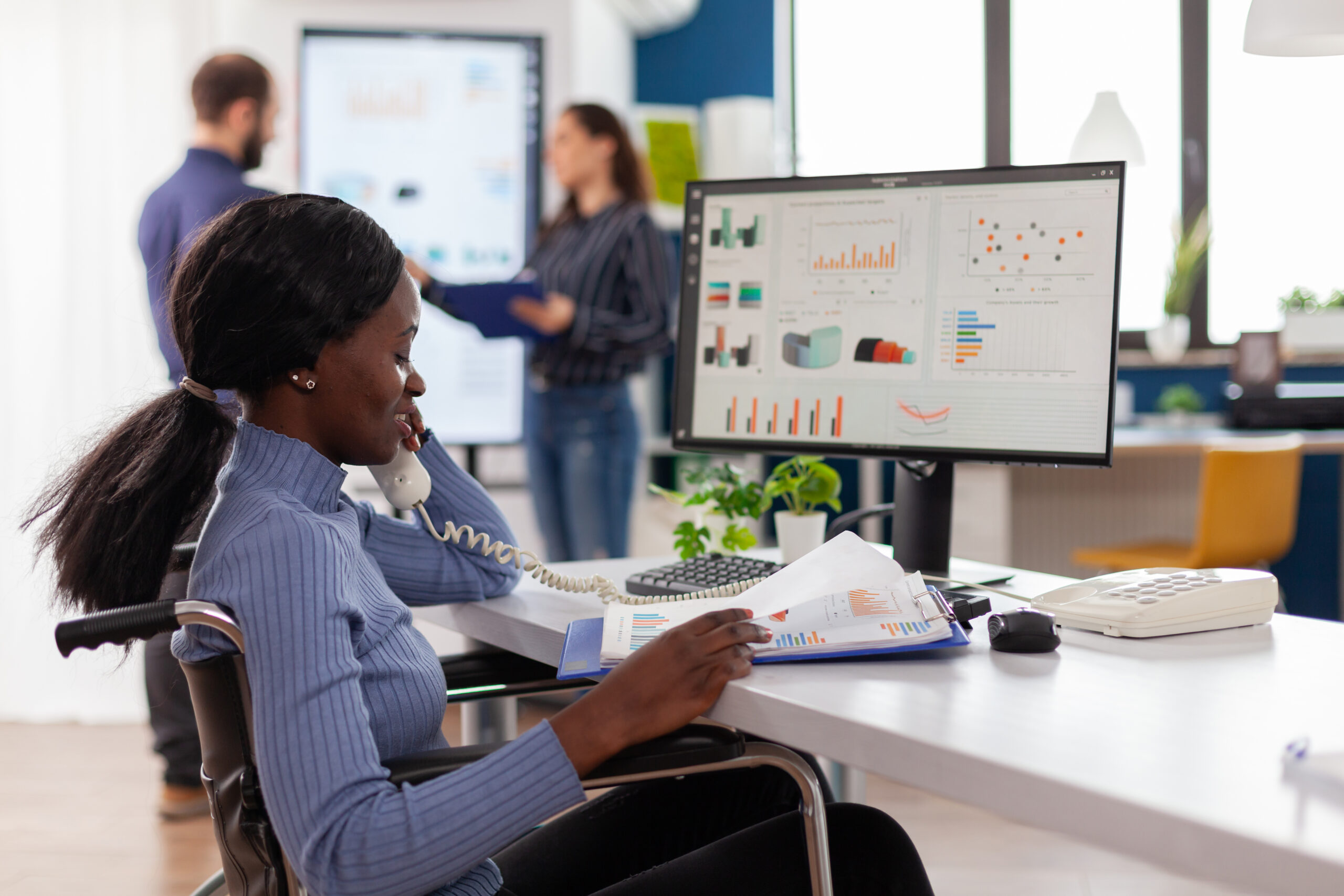 Busy paralysed handicapped black employee answering phone talking with colleague sitting immobilized in wheelchair in start up business office resolving financial problems discussing statistics graphs.